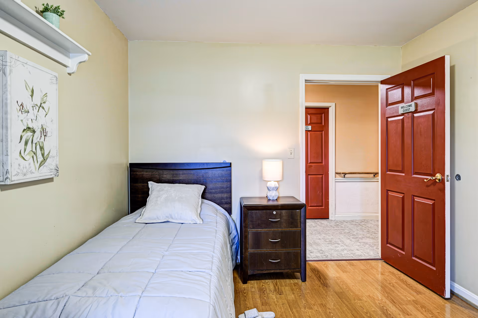 A small bedroom with a single bed covered in a light gray comforter and a white pillow. Next to the bed is a dark wooden nightstand with three drawers and a white lamp on top. The walls are light-colored, and there is a floral painting hanging on the left wall. The door to the room is open, painted red with a sign that reads 'WELCOME GUEST'. Another red door is visible through the doorway.
