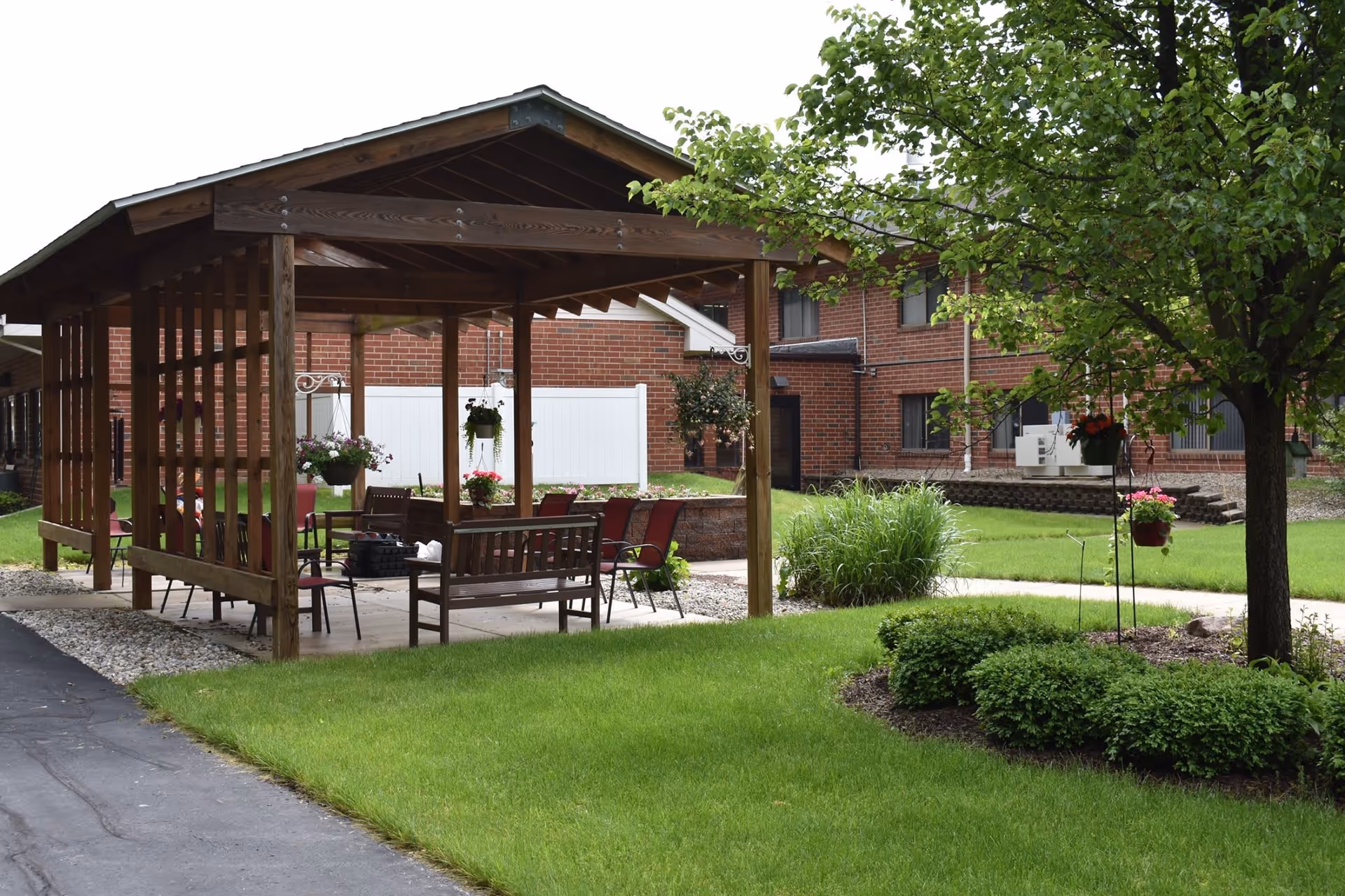 Outdoor covered seating area with wooden benches and chairs surrounded by green grass, bushes, and a tree. The background shows a brick building with windows and a white fence.