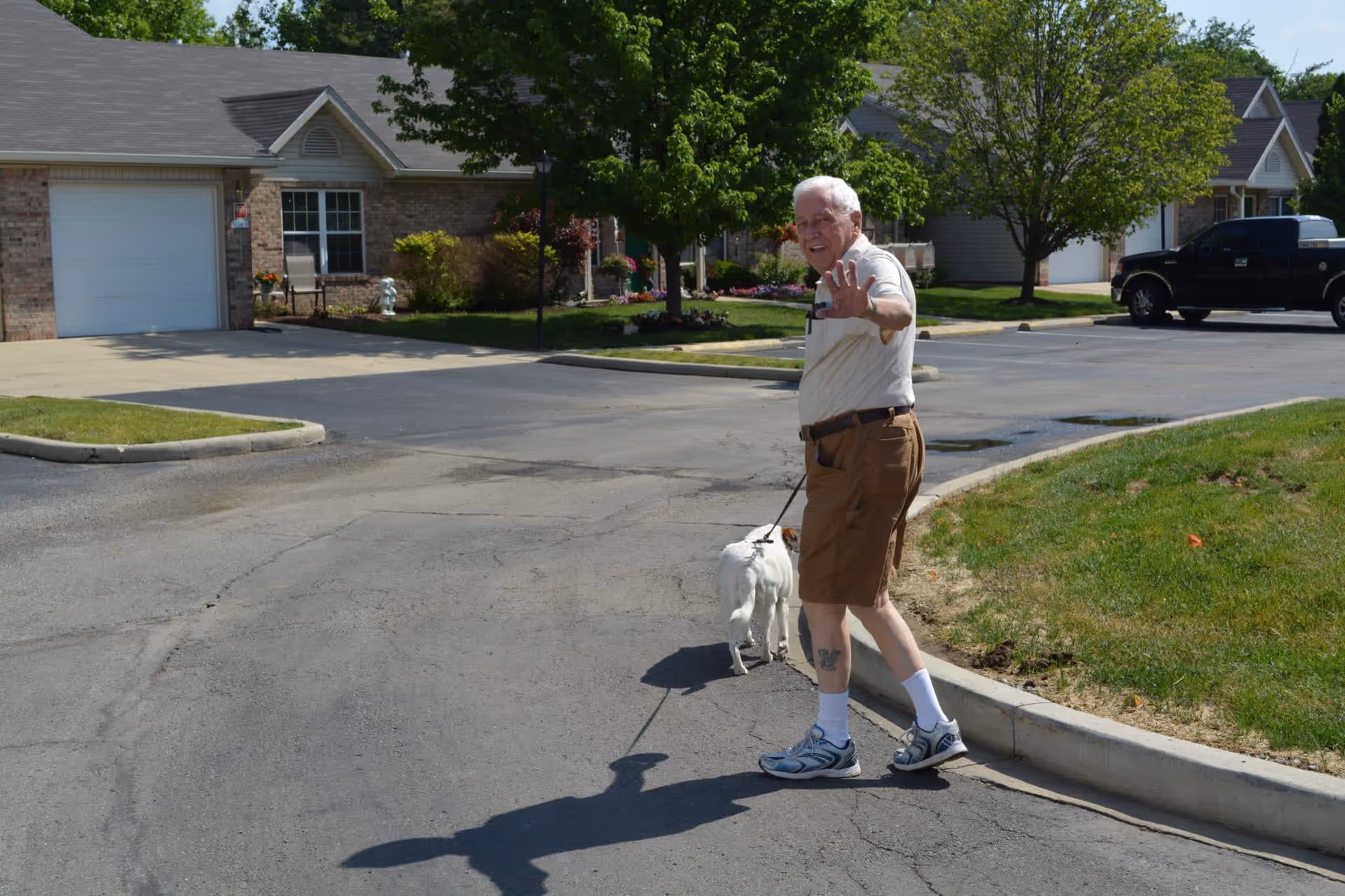 An elderly man wearing a light-colored shirt, brown shorts, white socks, and sneakers is walking a small white dog on a leash in a residential area with single-story houses, green lawns, and trees. The man is looking back and waving at the camera.