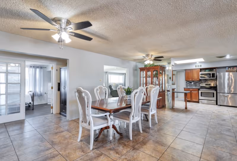 Spacious interior of an assisted living facility featuring a dining area with a wooden table and six white cushioned chairs, ceiling fans with lights, tiled floor, and an open kitchen with stainless steel appliances and wooden cabinets. A bedroom is visible through a doorway on the left.