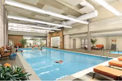 Indoor swimming pool area with clear blue water, surrounded by lounge chairs and plants. The space has a high ceiling with exposed beams and large windows letting in natural light. There are a few people swimming and relaxing by the pool.