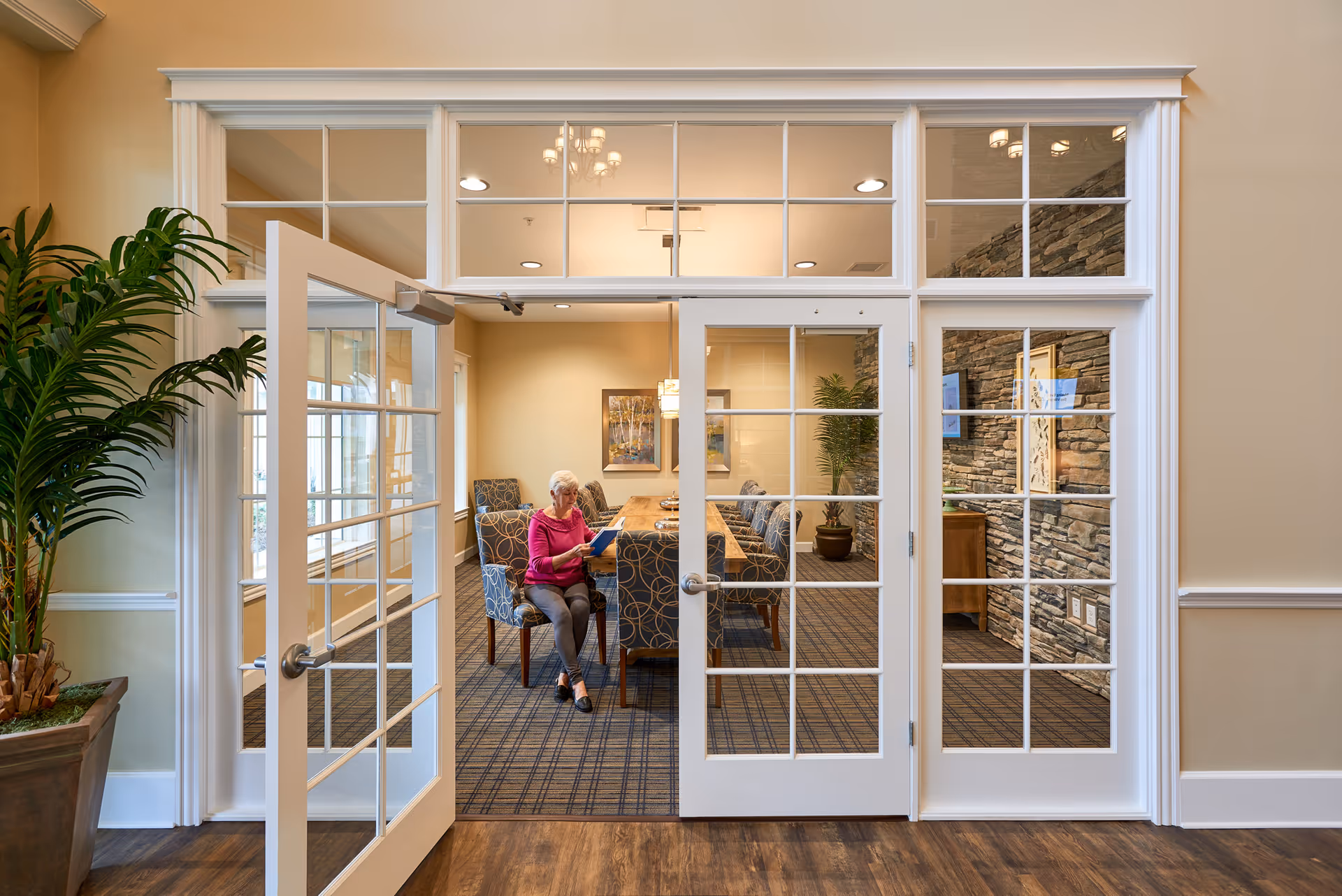 A senior woman sitting on a chair inside a well-lit meeting or dining room with large glass-paneled double doors open. The room features a long wooden table surrounded by patterned chairs, a stone accent wall, framed artwork, and potted plants.
