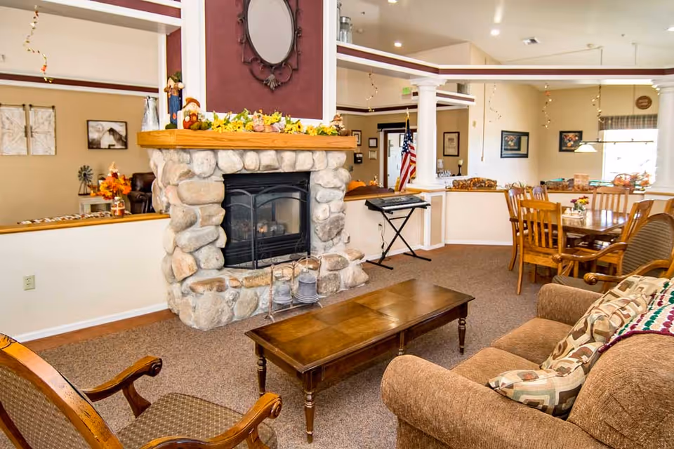 A cozy senior living facility common area featuring a stone fireplace decorated with autumn-themed garland and figurines. The room includes a brown sofa with patterned pillows, a wooden coffee table, a wooden dining table with chairs, and a keyboard on a stand near white columns. The walls are adorned with framed pictures and an American flag is visible in the background.
