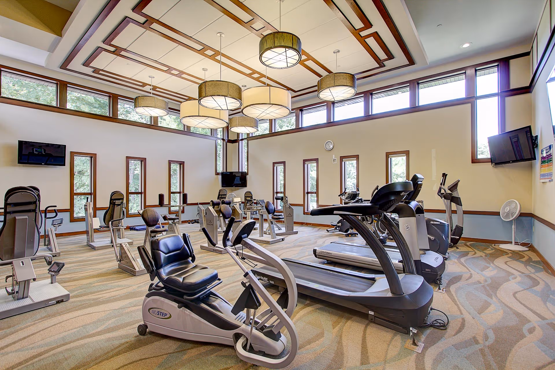 Bright, spacious fitness room with treadmills, recumbent bikes, and other exercise equipment under large pendant lights.