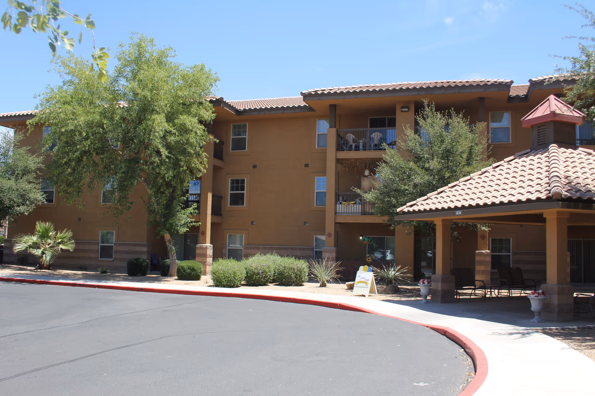 Exterior view of a multi-story senior living facility building with a covered seating area, trees, shrubs, and a clear blue sky.