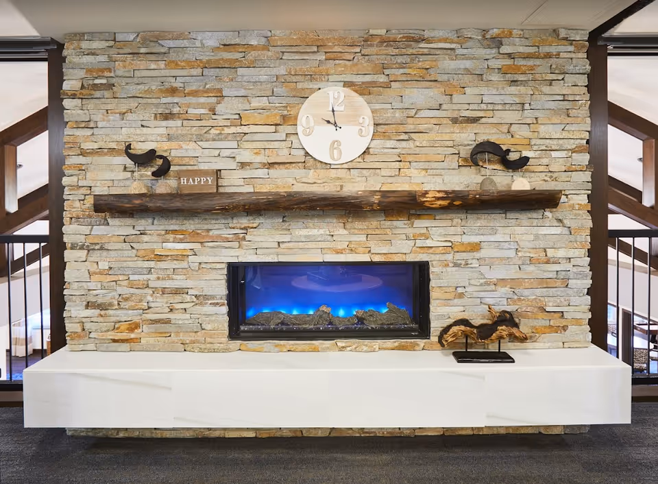 A modern stone fireplace with a wooden mantel displaying decorative items including a small sign that says 'HAPPY' and two bird sculptures. Above the mantel is a round clock showing the time as 12:15. The fireplace has a blue flame effect and is set in a stone wall with a white base below it. The surrounding area shows parts of a room with wooden beams and railings.