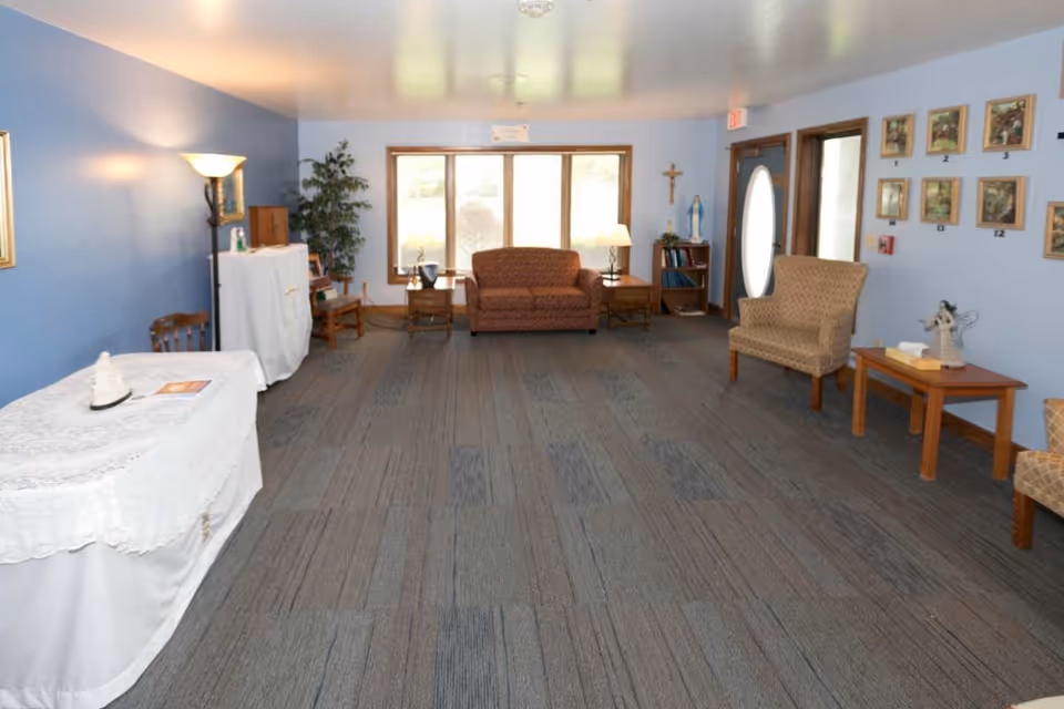 A spacious room with blue walls and carpeted floor featuring a small brown couch, two armchairs, wooden side tables with lamps, a bookshelf with religious statues, framed pictures on the wall, and a table covered with a white cloth. There is a large window and a door with an oval glass panel.