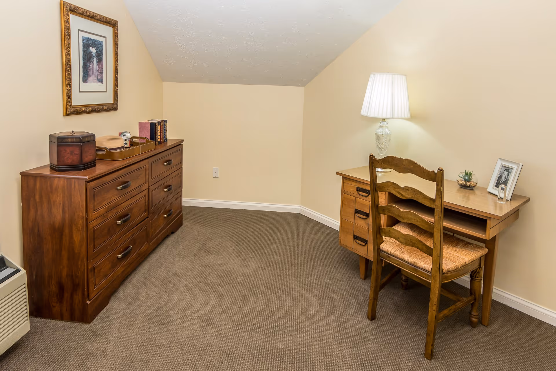 A small room with beige walls and carpeted floor featuring a wooden dresser with decorative items and books on top, a framed picture hanging above it, and a wooden desk with a chair, a table lamp, a small plant, and a framed photo on it.