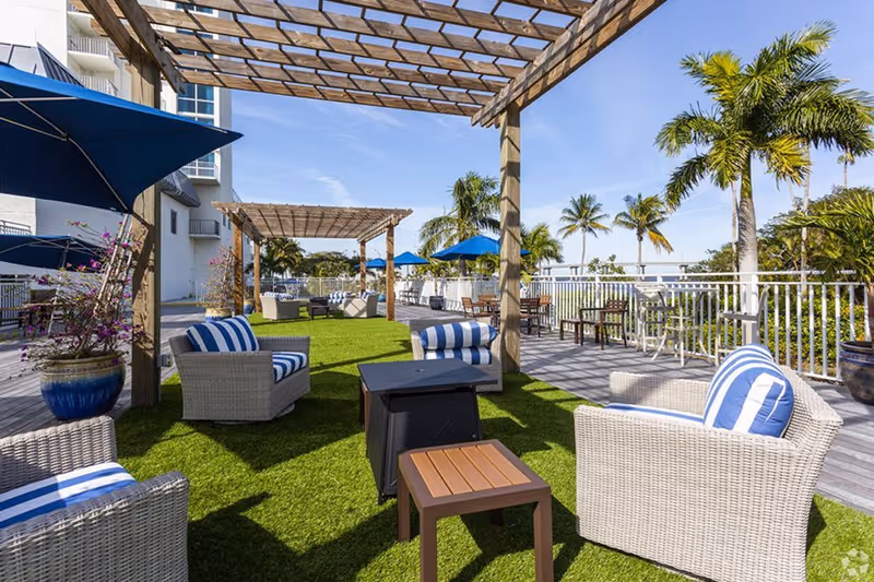Outdoor patio area with wicker chairs featuring blue and white striped cushions, wooden pergolas providing partial shade, potted plants, and palm trees under a clear blue sky.