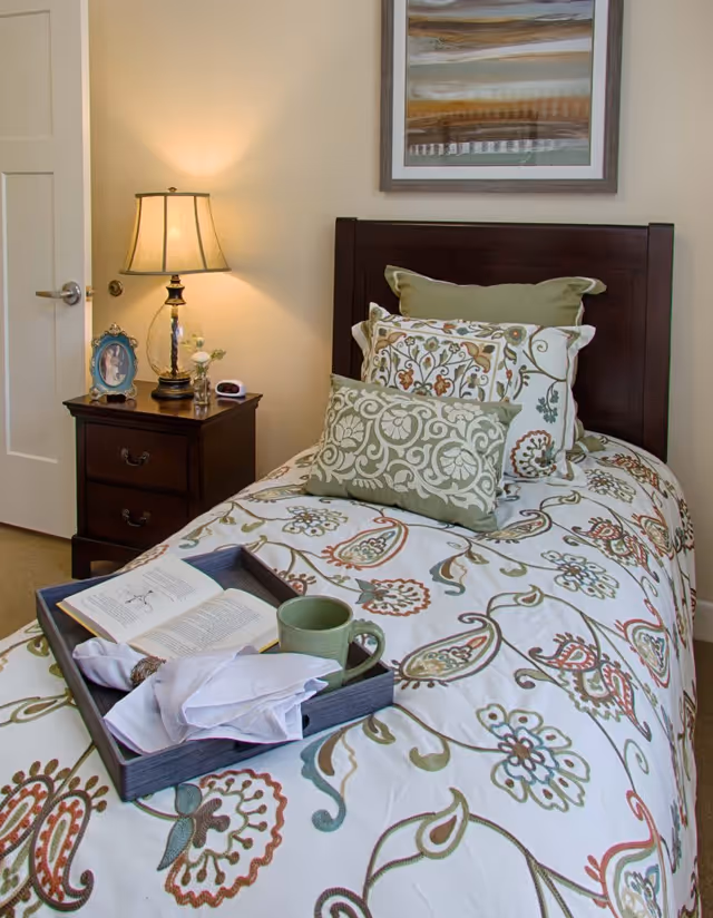 A neatly made single bed with a floral patterned bedspread and multiple decorative pillows. On the bed is a wooden tray holding an open book, a green mug, and a folded white napkin. Next to the bed is a dark wooden nightstand with a lamp, a framed photo, and a small alarm clock. A framed abstract painting hangs on the wall above the bed.