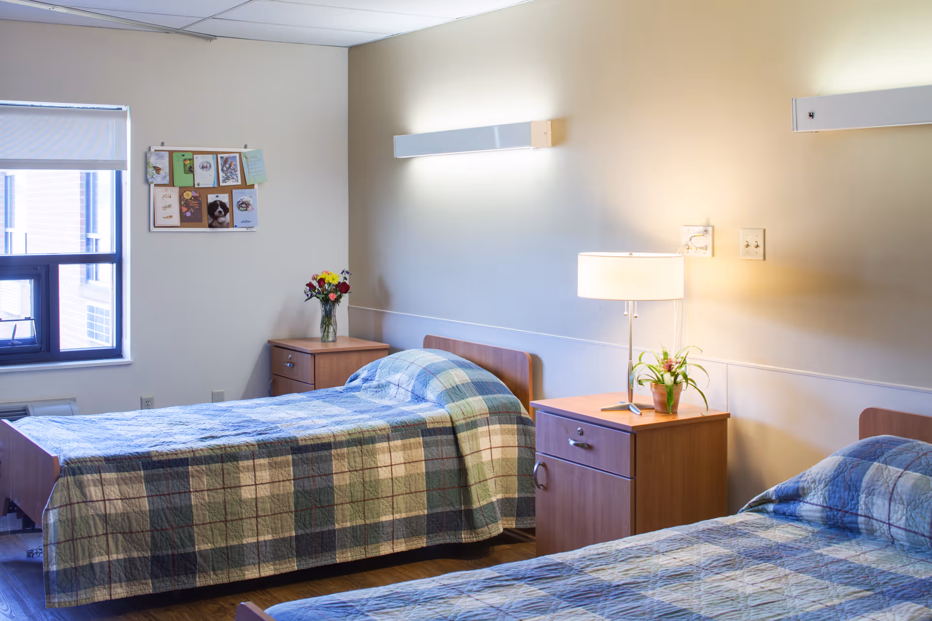 A bright room with two single beds covered in blue and green plaid quilts. Each bed has a wooden headboard and is accompanied by a wooden nightstand. One nightstand has a vase with flowers and the other has a lamp and a potted plant. A window with a white blind is on the left wall, and a bulletin board with cards and pictures is mounted above the nightstand. The walls are painted a light beige color.