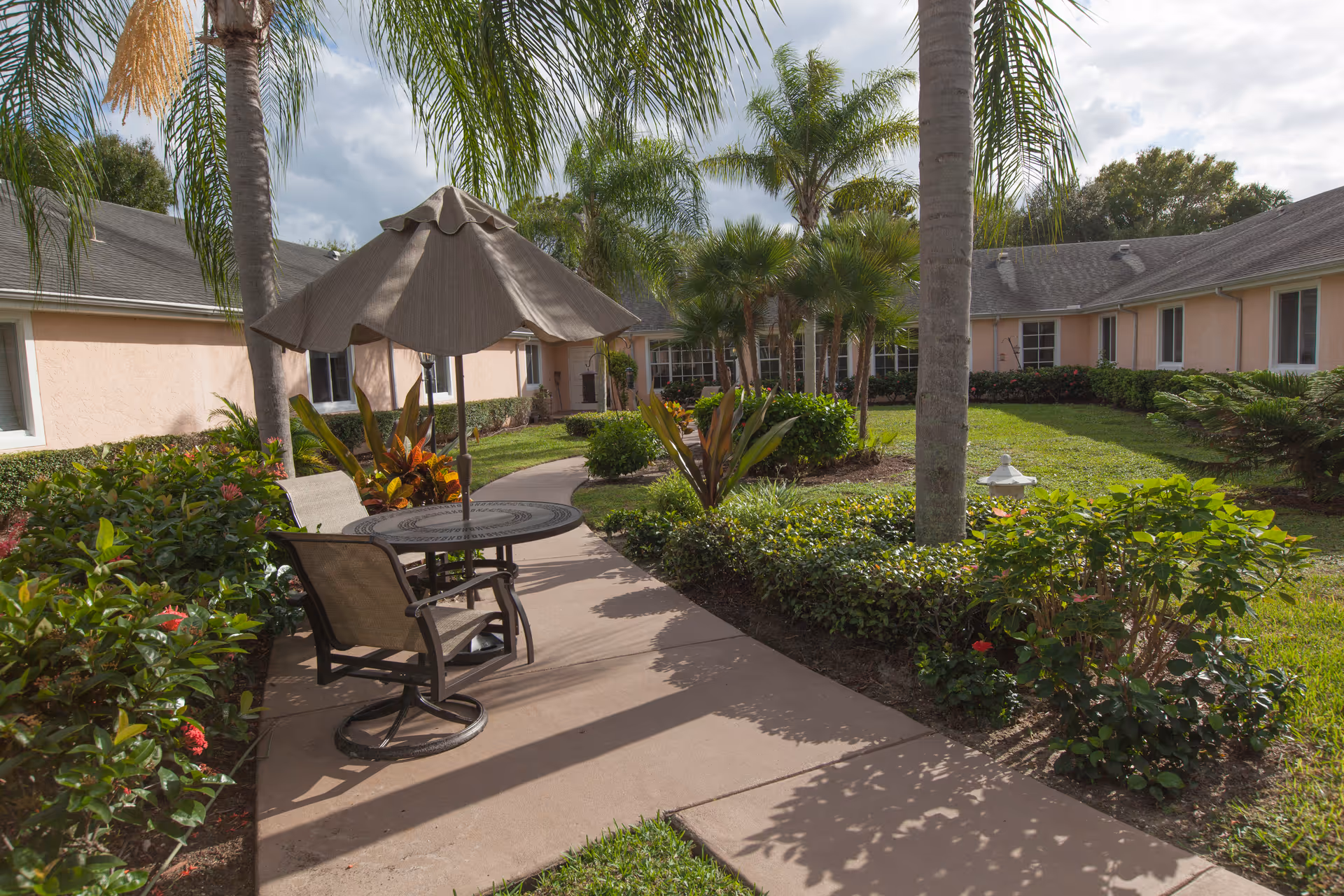 A sunny courtyard with a round patio table and umbrella beside a walkway, surrounded by palm trees, shrubs, and a single-story building.
