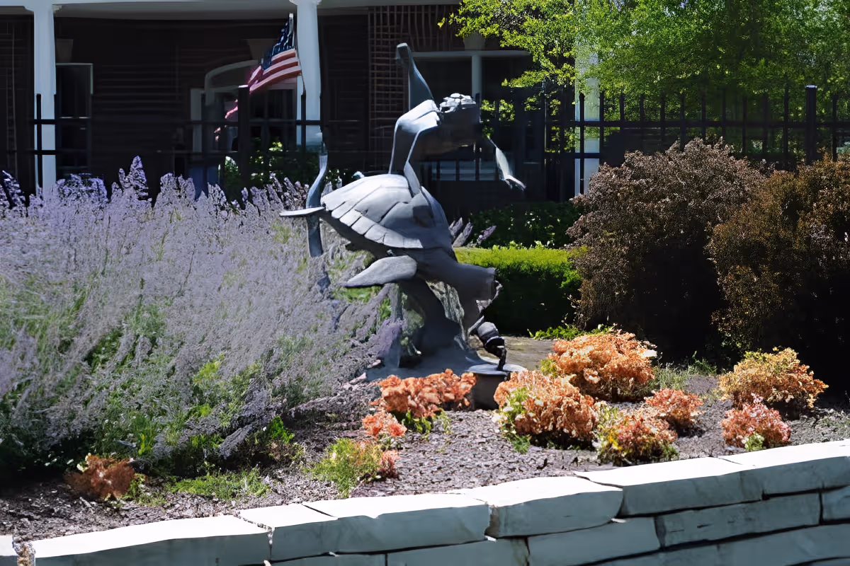 Bronze turtle sculpture in a landscaped garden bed with lavender, shrubs, and an American flag visible by a building.