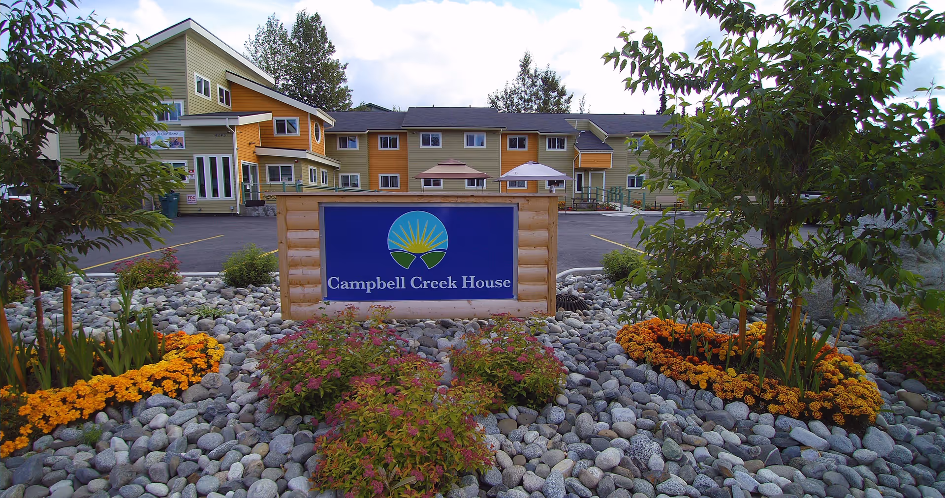 A wooden sign reading "Campbell Creek House" sits in a rock garden with flowers and small trees in front of a multi-story yellow-and-green building.