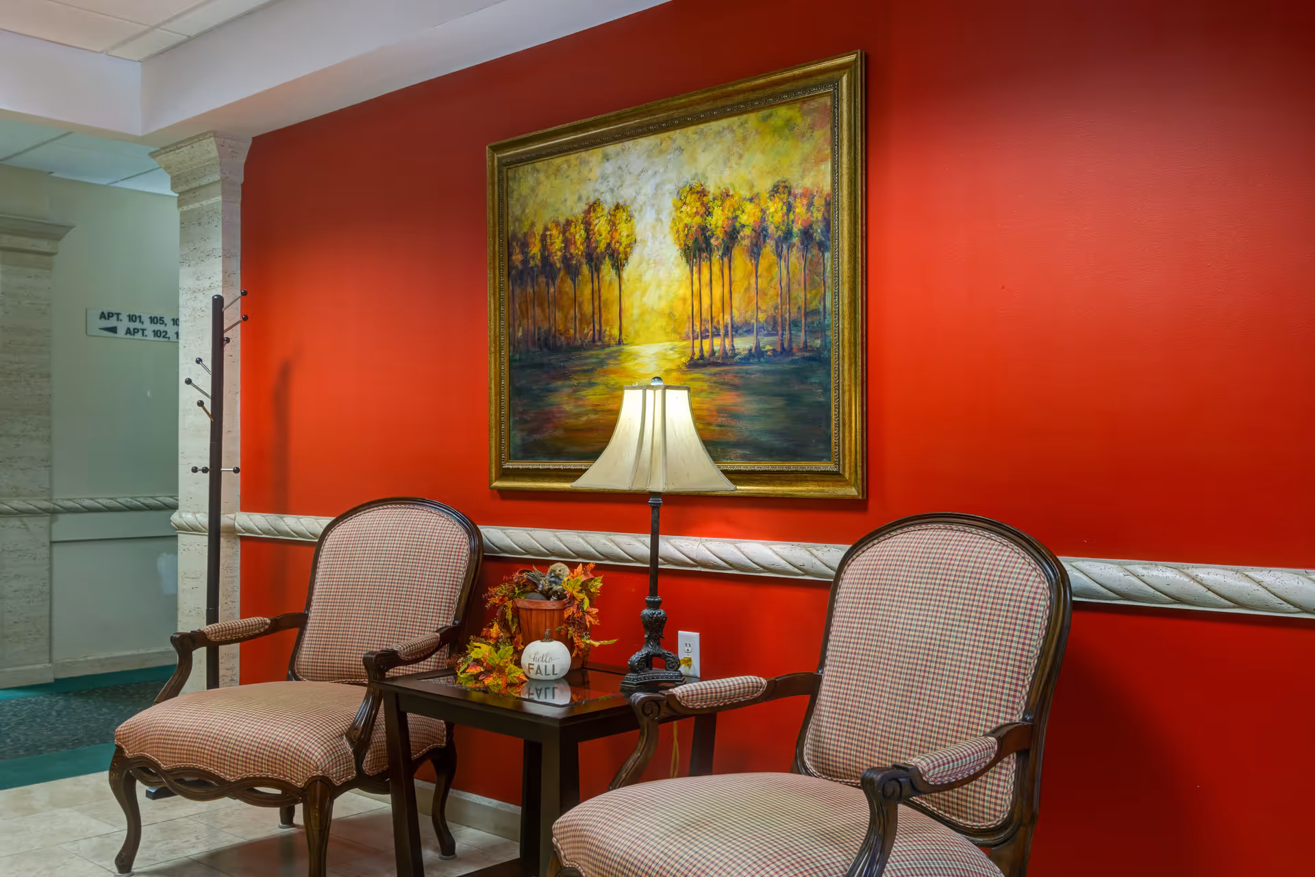 Two upholstered chairs flank a small table with a lamp and fall decorations beneath a framed painting on a red wall in a facility seating area.