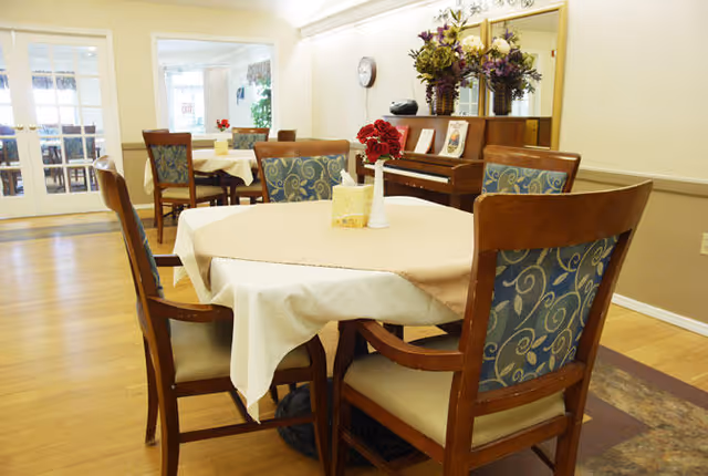 A dining area with wooden chairs featuring patterned blue cushions around tables covered with beige tablecloths. A piano with flower arrangements and framed pictures is visible against the wall, and a clock hangs above it. The room has wooden flooring and light-colored walls.