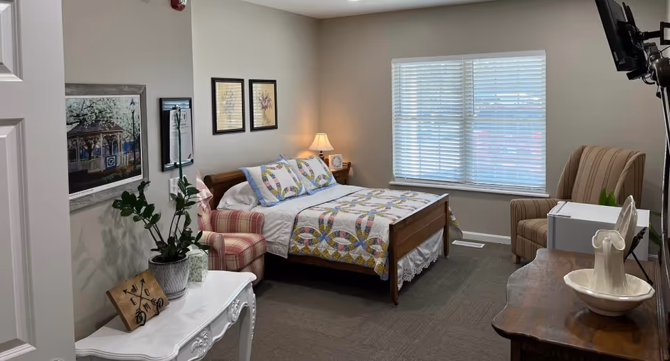 A cozy bedroom in an assisted living facility featuring a wooden bed with a colorful quilt and pillows, a striped armchair, a window with blinds, a small white table with a plant and decorative items, framed artwork on the walls, and a TV mounted on the wall.