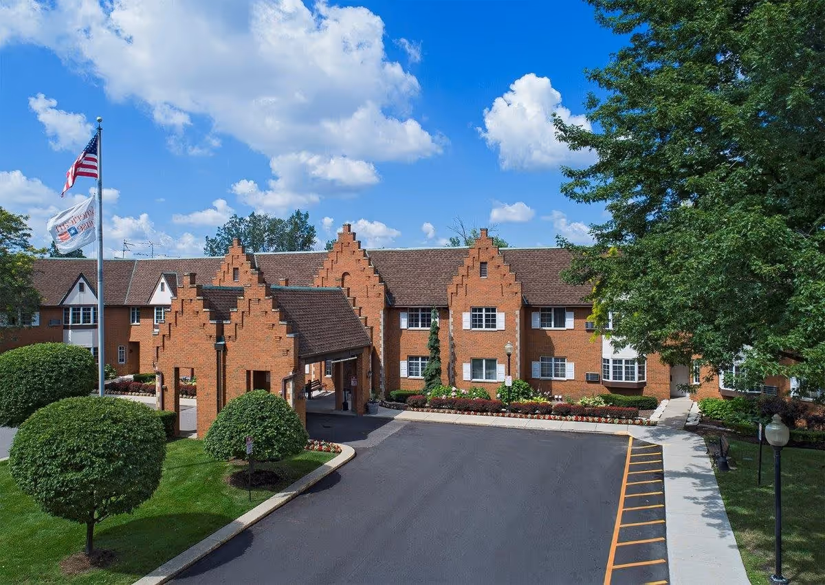 Exterior view of a brick senior living facility building with a peaked roof design, a covered entrance, neatly trimmed bushes, a flagpole with the American flag and another flag, and a clear blue sky with some clouds.
