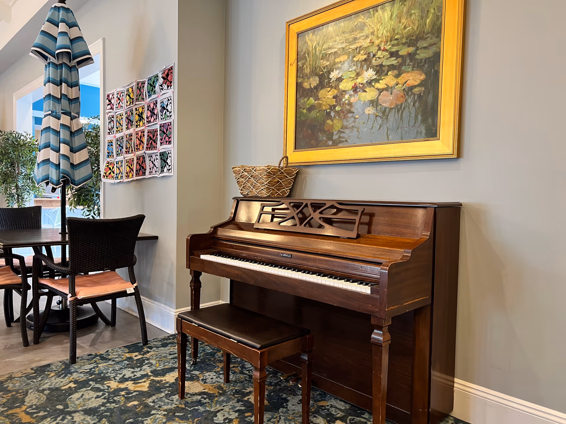 A cozy interior corner featuring a wooden upright piano with a matching bench, a woven basket on top, and a framed painting of water lilies hanging above. To the left, there is a dining table with four wicker chairs and a closed blue and white striped umbrella. A colorful quilt or fabric art piece is displayed on the wall behind the table, and some green plants are visible in the background.