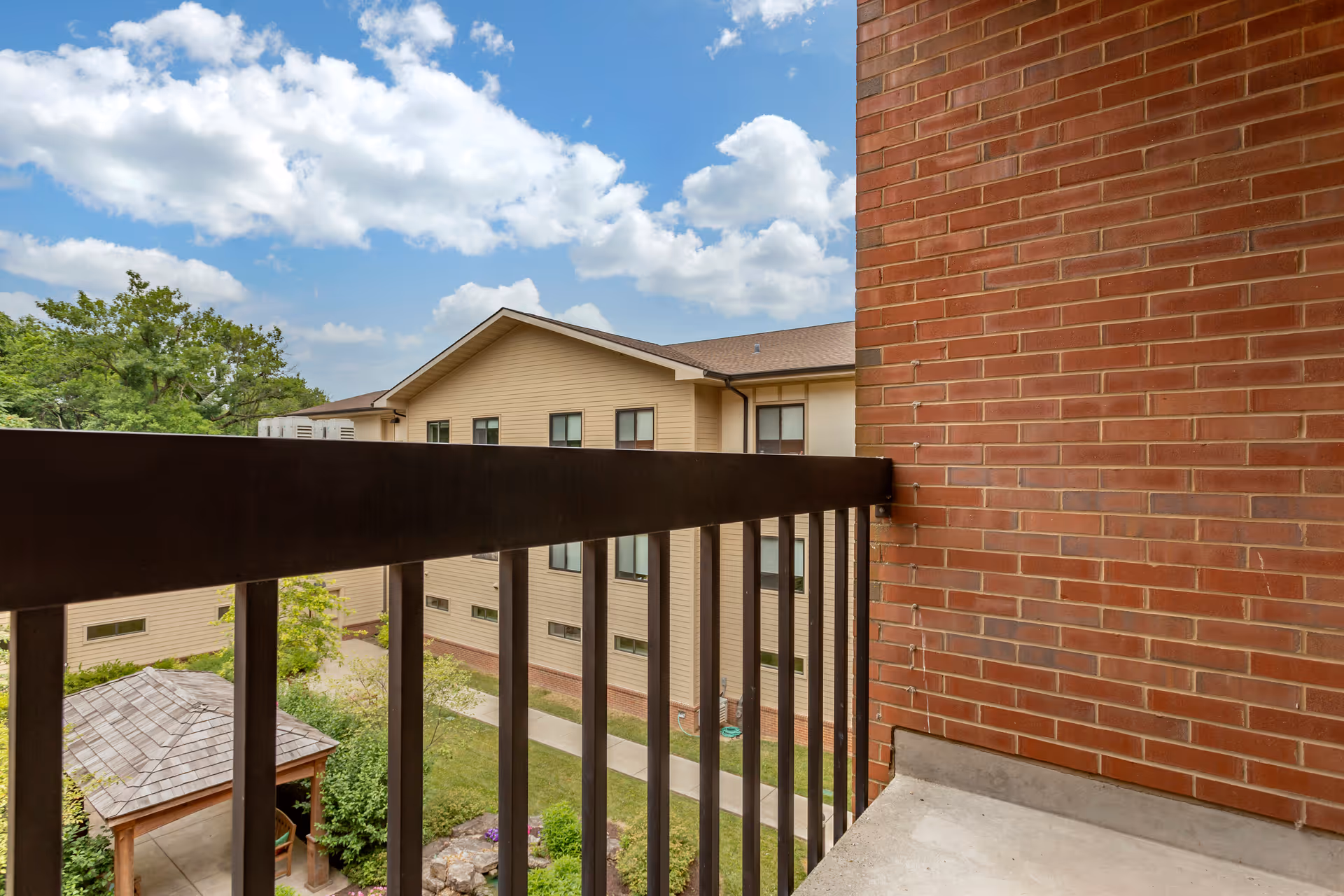 View from a balcony with black metal railing overlooking a courtyard with a wooden gazebo, green trees, and a beige multi-story building under a partly cloudy blue sky.
