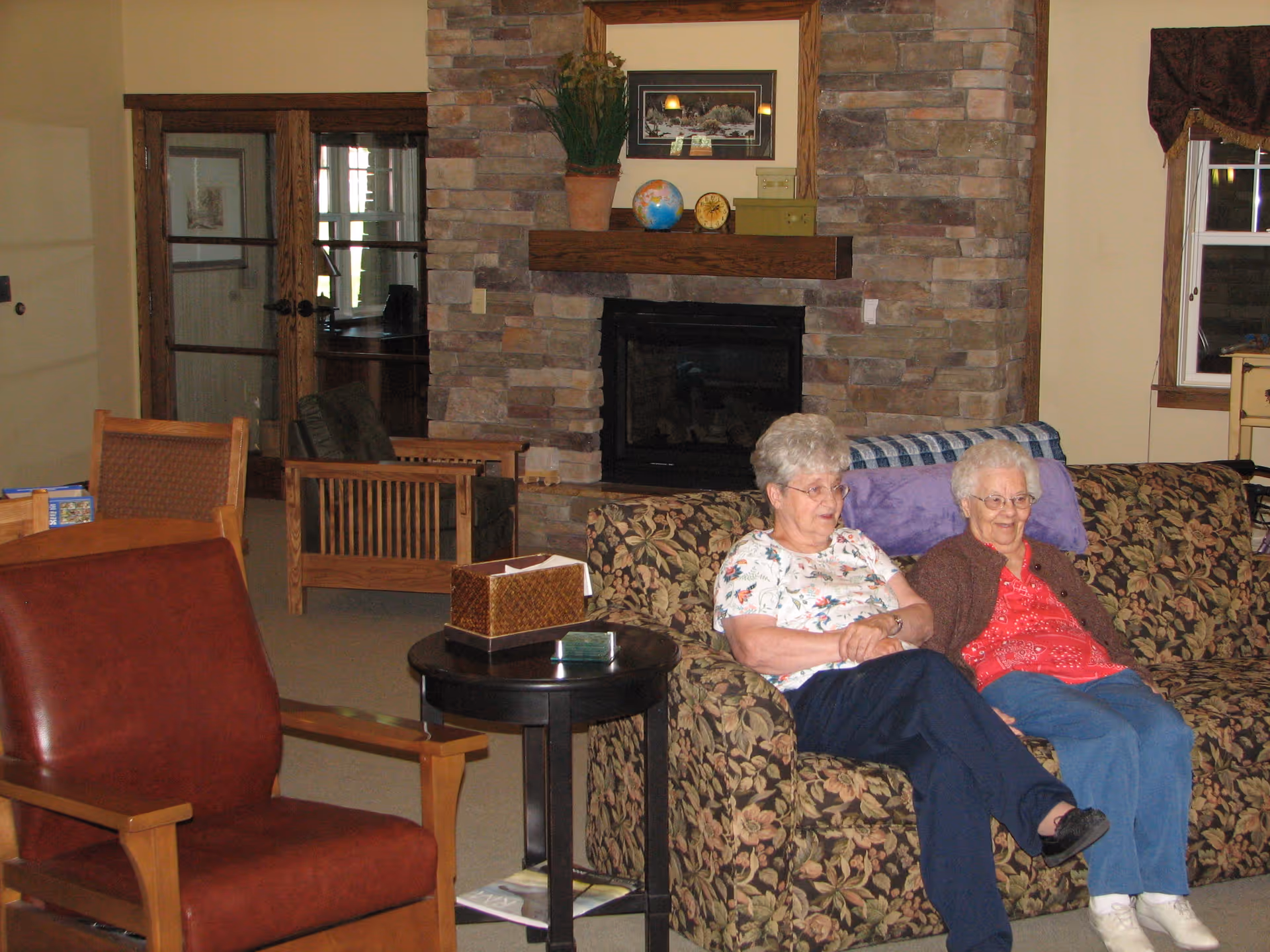 Two elderly women sitting on a floral patterned couch in a cozy living room with a stone fireplace behind them. The room has wooden furniture, a small round table with a tissue box, and a window with curtains.