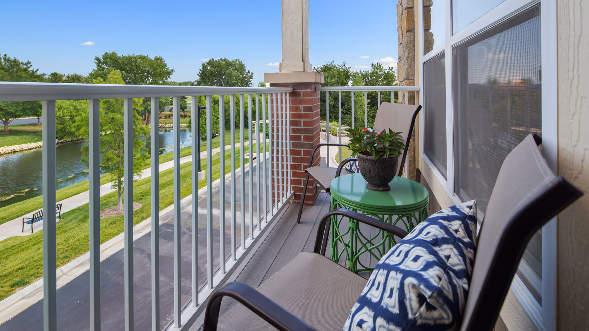 Balcony with two beige chairs, one with a blue and white patterned pillow, and a green round table with a potted plant. The balcony overlooks a small river, green grass, trees, and a walking path under a clear blue sky.