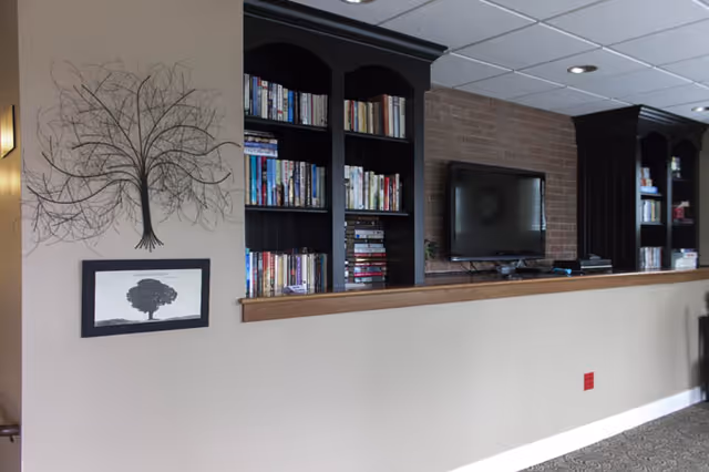 Interior view of a room with a beige half-wall topped with a wooden ledge. Behind the wall are built-in black bookshelves filled with books and a flat-screen TV mounted on a brick wall. On the left side of the image, there is a metal wall art piece shaped like a tree and a framed picture of a tree below it. The ceiling has recessed lighting.