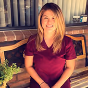 A woman with blonde hair wearing a maroon scrub top is sitting on a wooden bench outside a building with a brick wall and window behind her. There is a small green plant on the bench next to her.