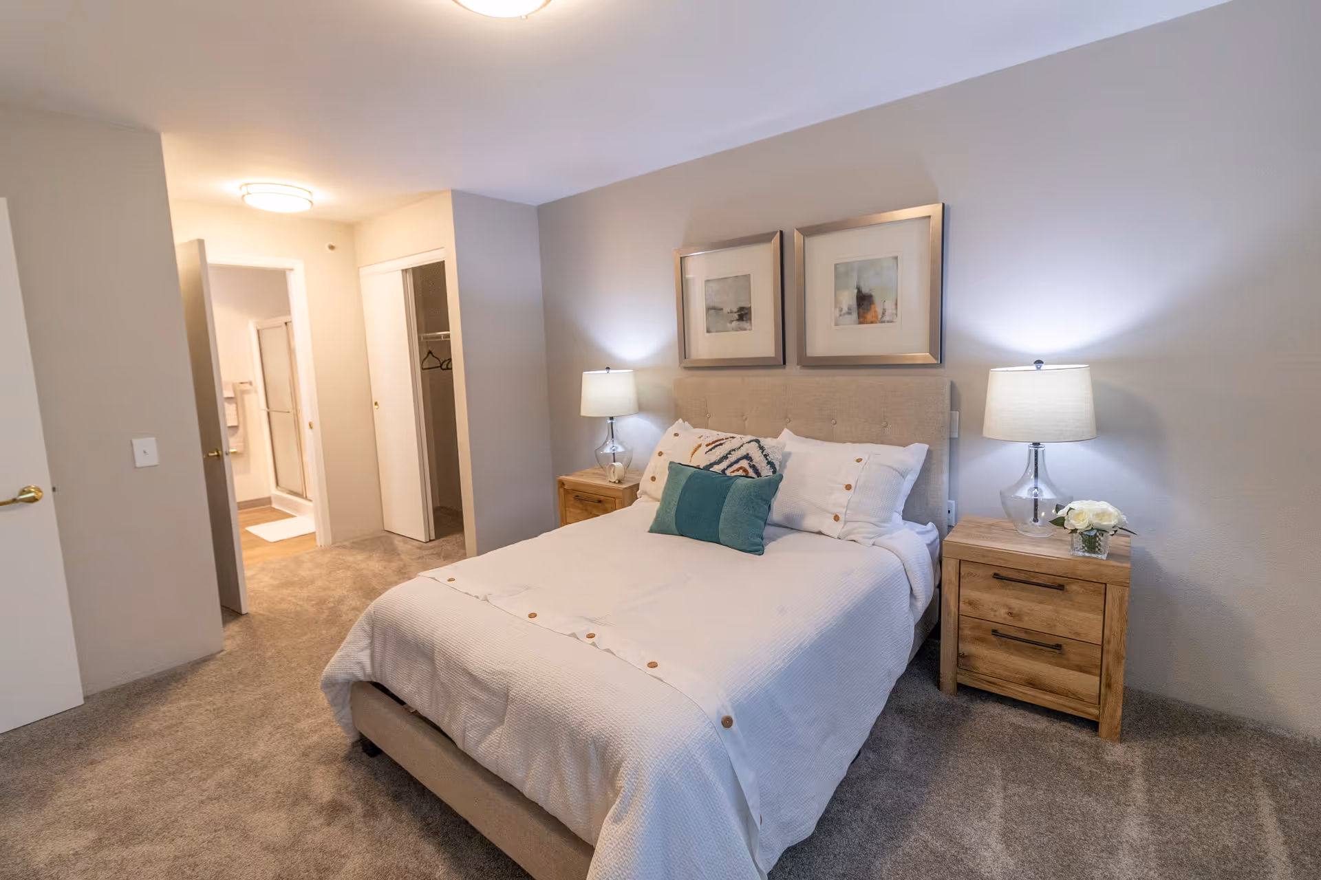 A cozy bedroom with a neatly made bed featuring white bedding and decorative pillows. There are two wooden nightstands on either side of the bed, each with a lamp and one with a small flower arrangement. The walls are painted light gray, and two framed artworks hang above the bed. In the background, there is an open door leading to a bathroom and a closet with sliding doors.