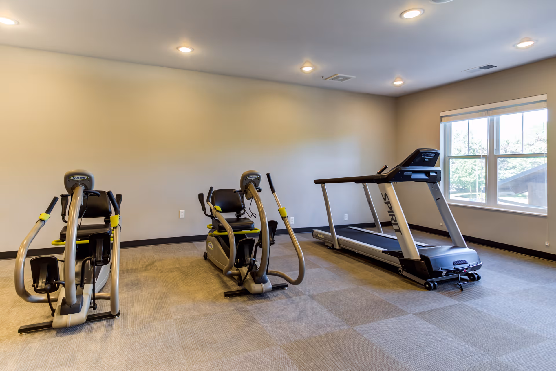 Small fitness room with two recumbent exercise bikes and a treadmill by a window.