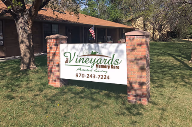 A sign for The Vineyards Memory Care assisted living facility mounted between two brick pillars on a grassy lawn with a tree and a building with a porch and an American flag in the background.