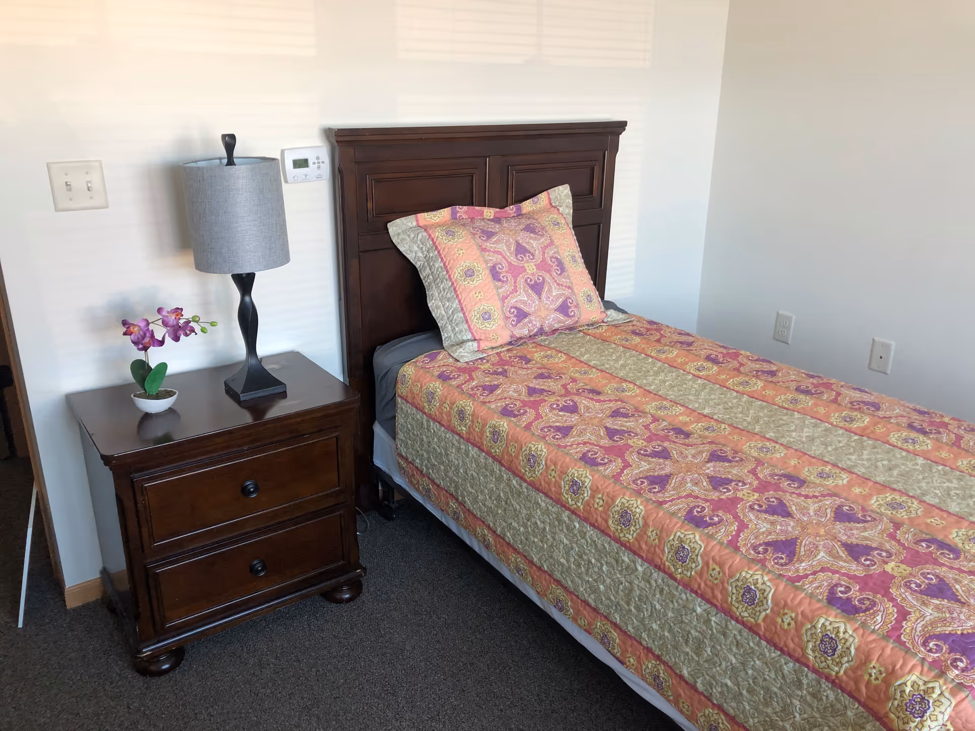 A single bed with a wooden headboard and a colorful patterned bedspread in shades of pink, purple, and green. Next to the bed is a wooden nightstand with two drawers, a gray table lamp, and a small potted purple orchid. The room has white walls and a carpeted floor.