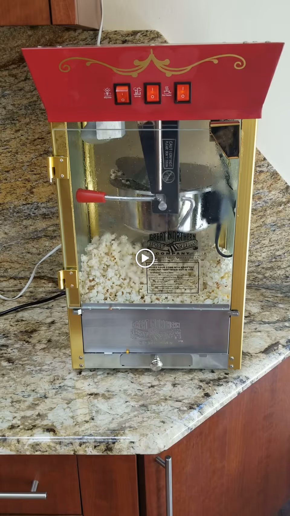A vintage-style popcorn machine with a red top and gold trim sits on a granite countertop. The machine is filled with freshly popped popcorn and has three red switches on the top front panel. The background shows a beige wall and wooden cabinets below the countertop.