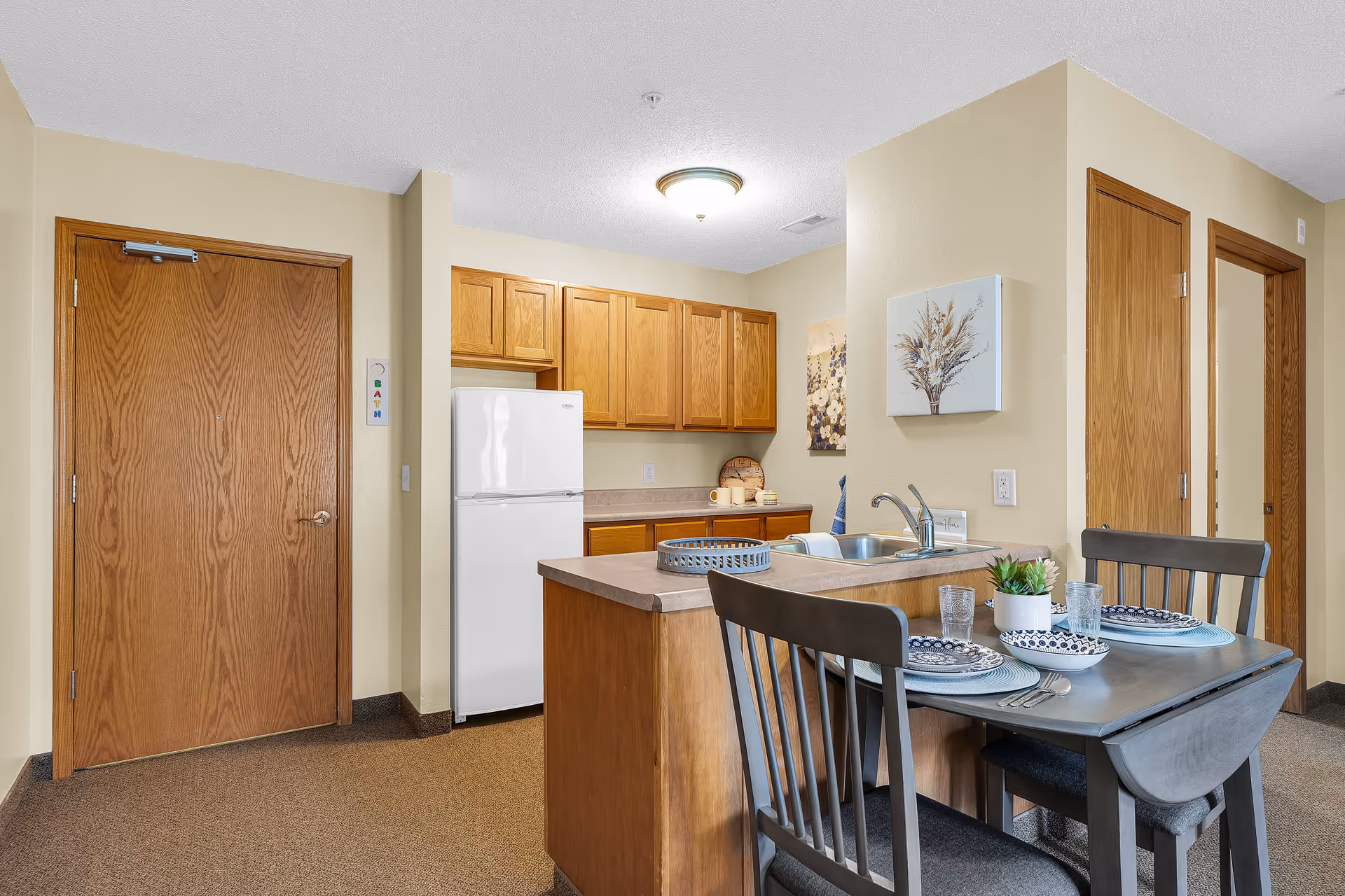 Interior view of a small kitchen and dining area in an assisted living facility. The kitchen has wooden cabinets, a white refrigerator, a countertop with a sink, and a small dining table set for two with plates, glasses, and a small plant. There are wooden doors and light beige walls in the background.