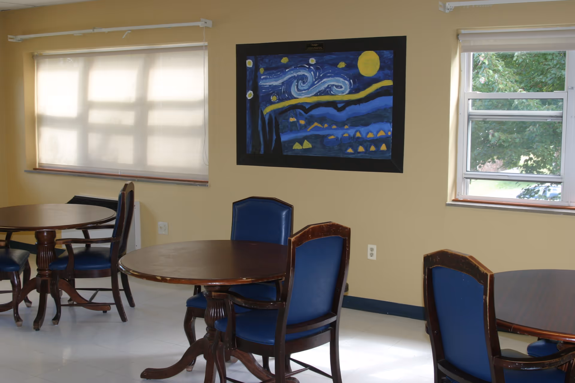 A room with round wooden tables and blue cushioned chairs. The walls are painted yellow, and there are two windows, one with a white blind and the other showing green trees outside. A colorful painting resembling Van Gogh's Starry Night is hung on the wall.