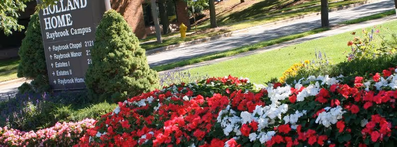 A landscaped garden area with vibrant red, white, and pink flowers in front of a sign that reads 'Holland Home Raybrook Campus' with directional information to various campus locations. The background shows a grassy area, trees, and a street with a yellow fire hydrant.