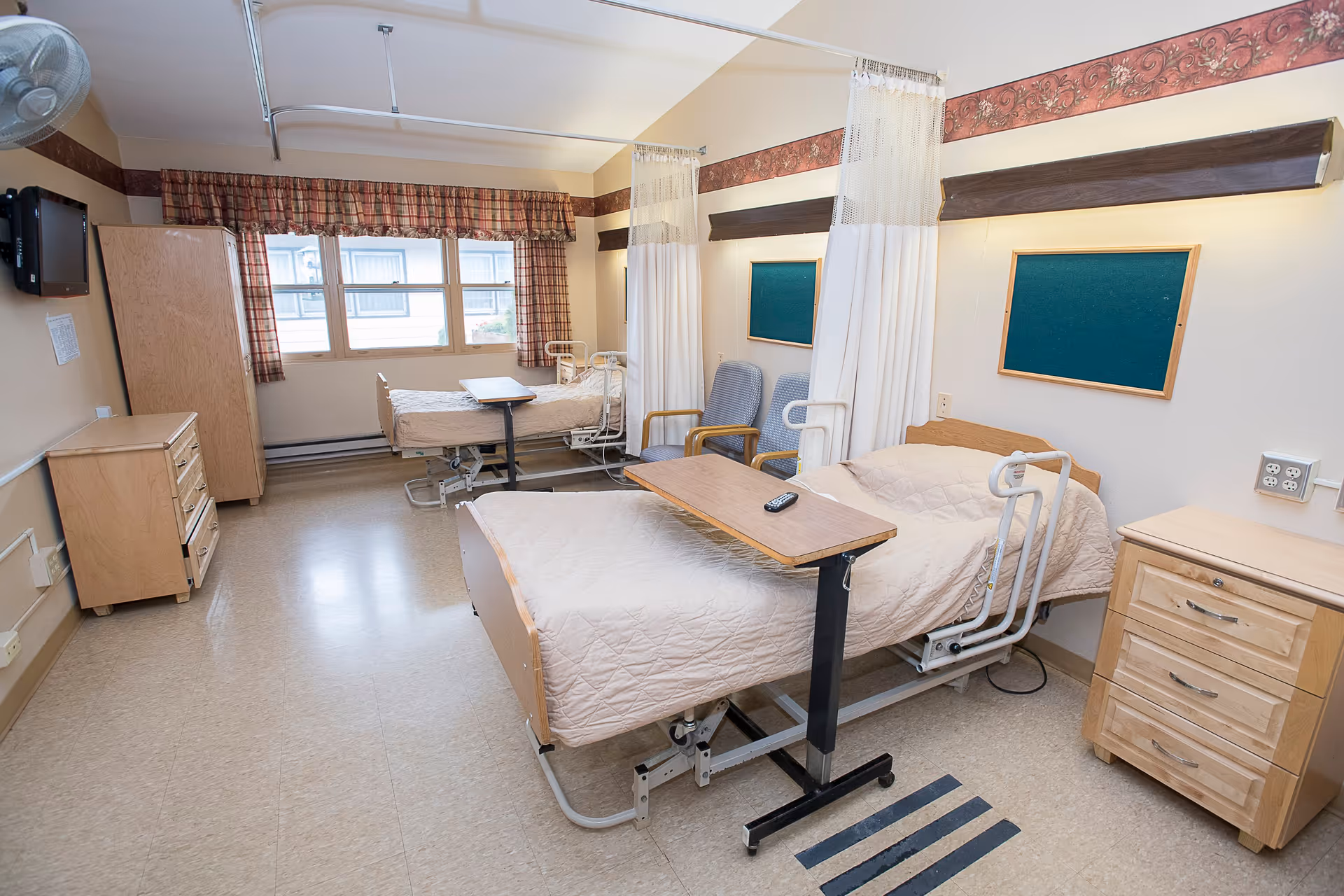 A shared patient room in a healthcare facility with two hospital beds, each equipped with a beige quilted blanket and an overbed table. The room has two chairs between the beds, privacy curtains, a window with plaid curtains, wooden dressers, and a wall-mounted TV.