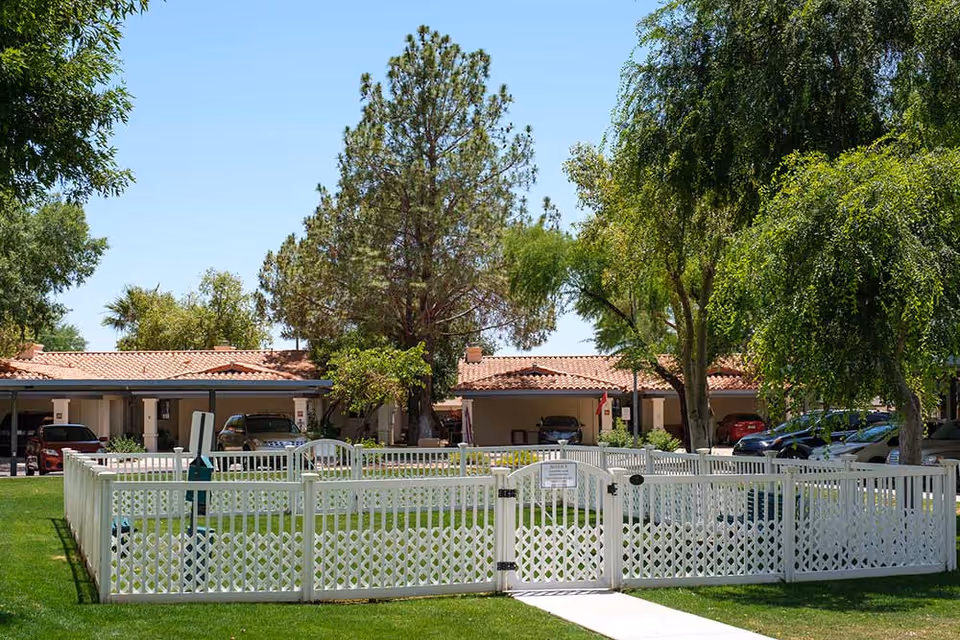 A fenced grassy area with a white lattice fence and gate in the foreground, surrounded by trees and greenery. Behind the fenced area are single-story buildings with red-tiled roofs and carports with parked cars. The sky is clear and blue.