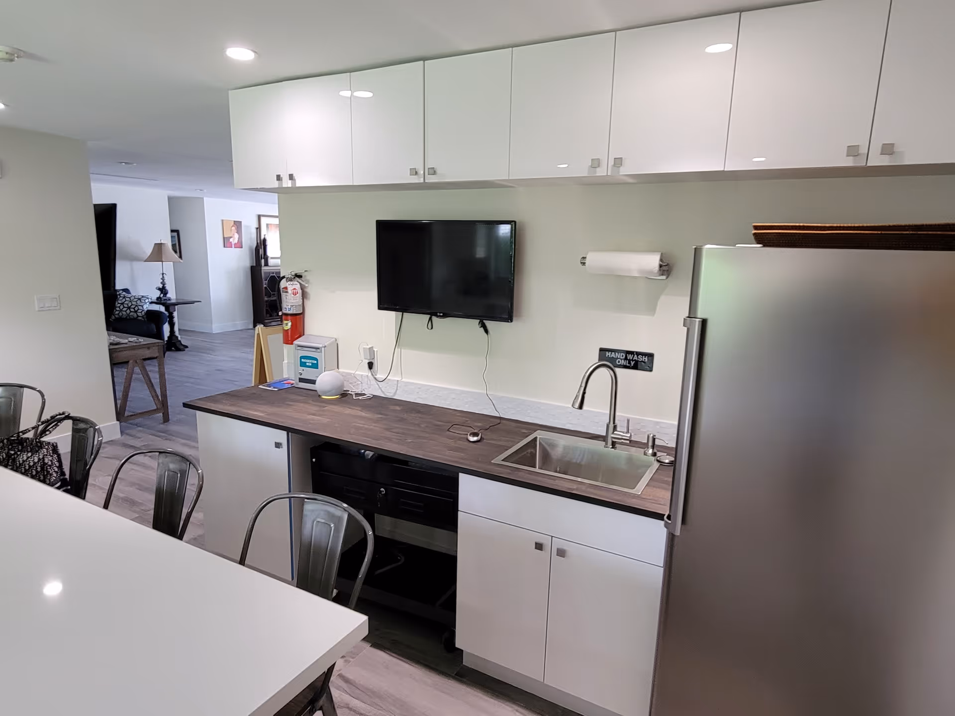 Modern kitchenette with a sink, stainless steel refrigerator, wall-mounted TV, white cabinets and countertop seating.