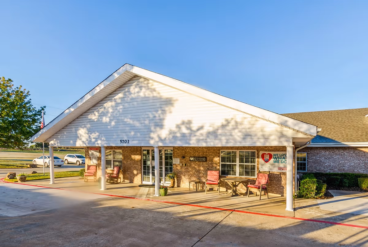 Front exterior view of a single-story brick building with a white gabled roof and a covered porch area. The porch has several chairs and a small table, with potted plants placed near the entrance. There is a sign on the wall that says 'welcome' and another sign with a heart logo and text. The building number 5701 is visible above the entrance. A few cars are parked in the background under a clear blue sky.