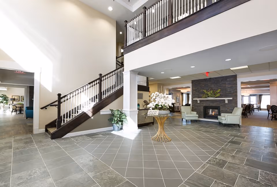 Bright and spacious senior living facility lobby with a staircase featuring dark wood handrails and white balusters. The floor is tiled with a mix of large gray tiles and smaller diamond-patterned tiles in the center. A round table with a floral arrangement sits in the middle of the room. To the right, there is a cozy seating area with two light green armchairs in front of a stone fireplace decorated with greenery. The area extends into a dining room with multiple tables and chairs visible in the background.