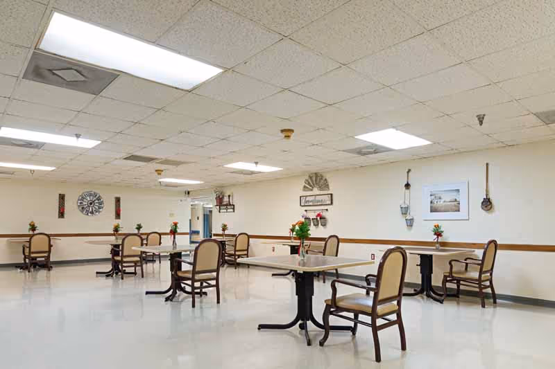 A spacious dining room with several tables and chairs arranged neatly. Each table has a small vase with flowers. The walls are decorated with various wall hangings and framed pictures. The ceiling has fluorescent lighting panels.