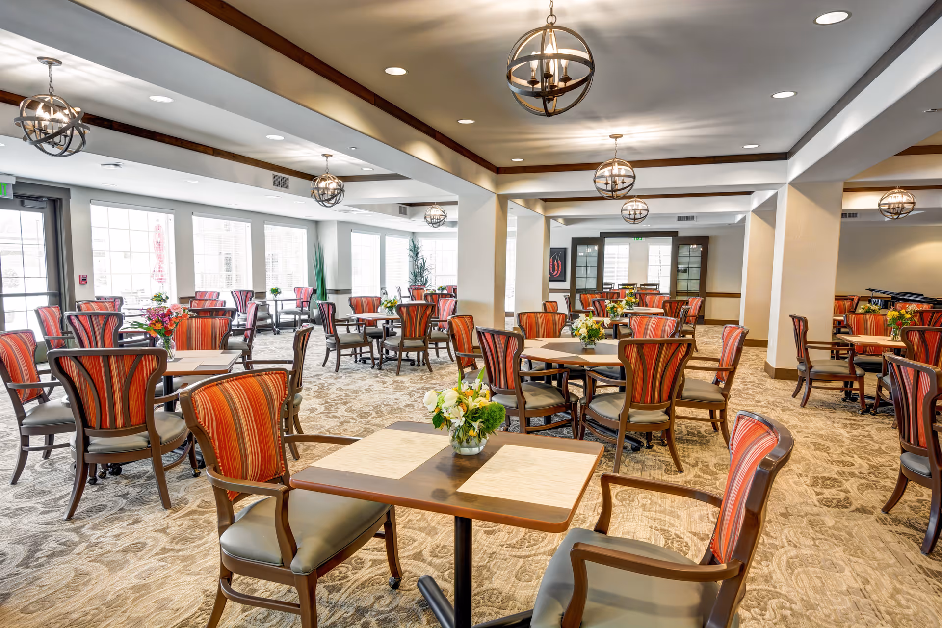 A spacious and well-lit dining room with multiple tables and chairs arranged neatly. The chairs have wooden frames with red and orange striped upholstery. Each table has a small floral centerpiece. The room features large windows allowing natural light to fill the space, and modern spherical pendant lights hang from the ceiling. The carpet has a patterned design, and the walls are painted in neutral tones.