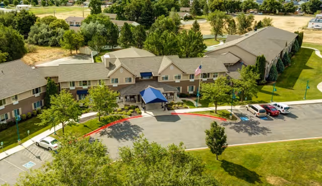 Aerial view of The Auberge at North Ogden senior living facility showing a large two-story building with a blue canopy entrance, surrounded by green trees and a parking area with several cars.