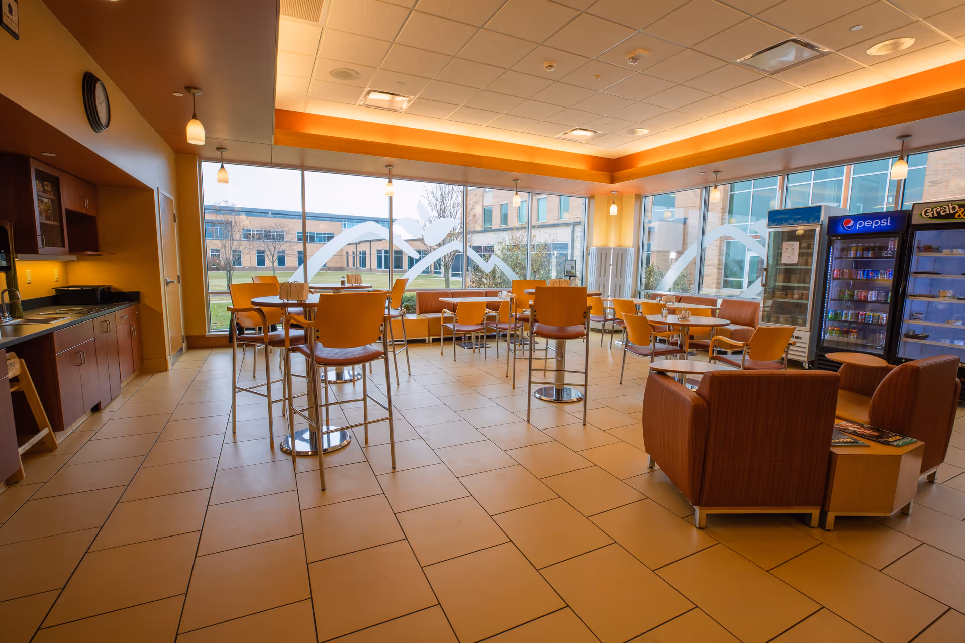 Bright communal dining and lounge area with tables, chairs, vending machines and large windows overlooking a courtyard.