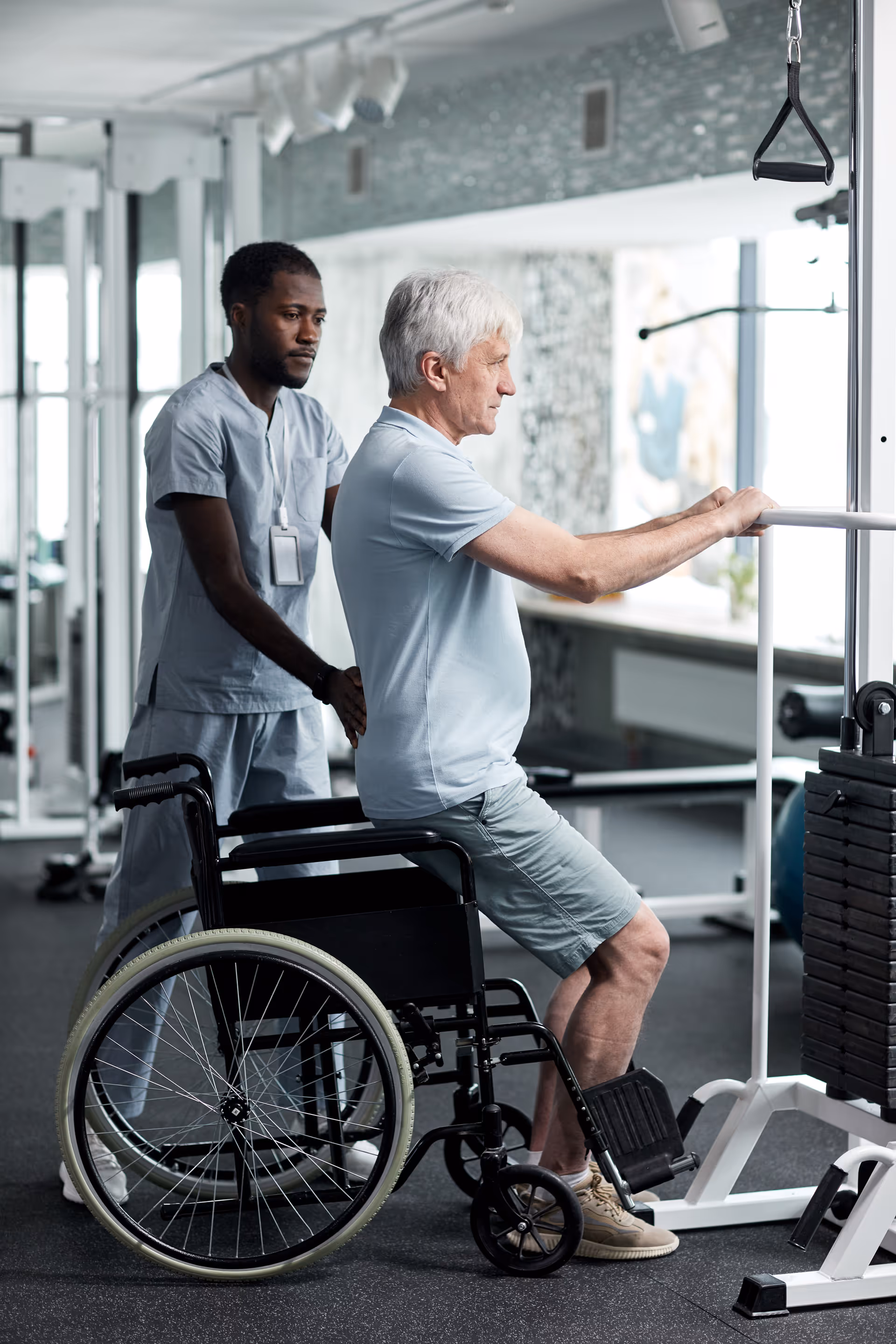 An elderly man in a wheelchair is assisted by a healthcare professional as he exercises using a parallel bar in a rehabilitation gym setting with exercise equipment in the background.