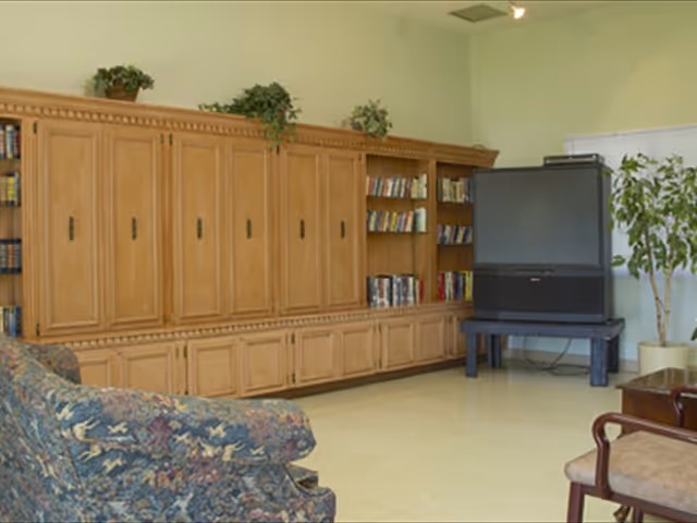 A living room area with a large wooden entertainment center filled with books, a large older-style television on a stand, a floral patterned sofa, a wooden chair with a cushioned seat, and a potted plant in the corner.