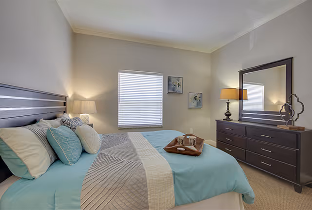 A neatly made bed with blue and beige bedding in a bedroom. The bed has multiple pillows and a wooden headboard. There is a wooden dresser with a large mirror and a lamp on top. Two framed floral artworks hang on the wall next to a window with closed blinds. The room is softly lit with neutral-colored walls and carpeted floor.