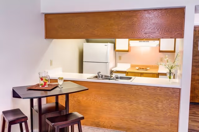 A small kitchen area with wooden cabinets, a white refrigerator, a stove with a range hood, and a sink set into a white countertop. In front of the kitchen counter is a small dark wooden table with two matching stools. On the table is a pitcher of fruit-infused water and a glass with a lemon slice. A vase with white flowers is placed on the kitchen counter.
