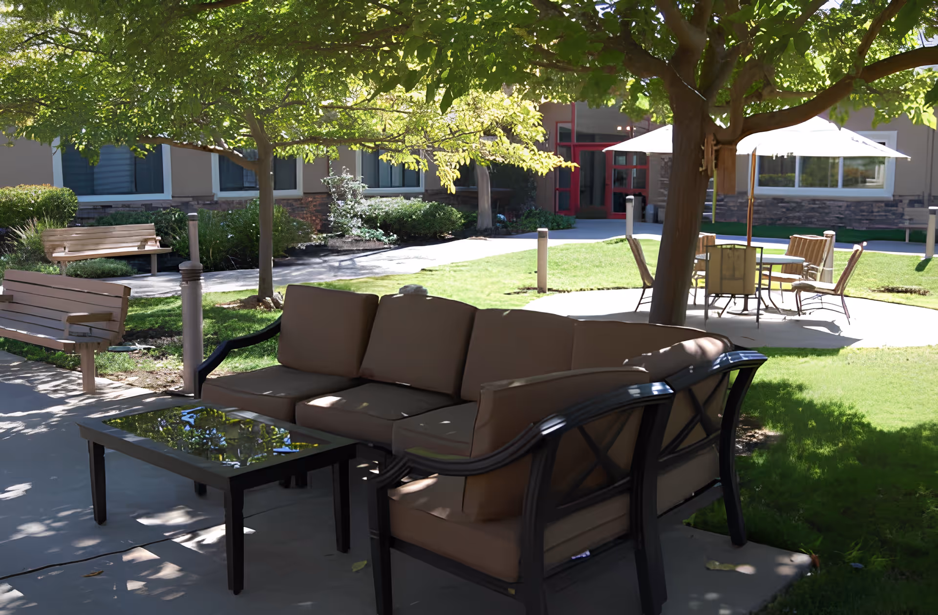 Outdoor seating area at Fair Oaks Estates with cushioned patio chairs and a glass-top table under a tree, surrounded by green grass, benches, and a building entrance in the background.